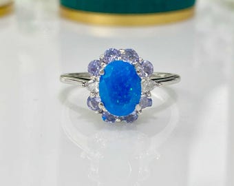 Può includere: Un anello d'argento con una grande pietra preziosa ovale blu brillante al centro. La pietra è circondata da pietre più piccole, viola chiaro e bianche, che creano un'aureola floreale. La fascia dell'anello è sottile e in argento lucido.