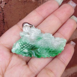 Pode incluir: Um pingente de jade verde esculpido em forma de drag&atilde;o com uma fivela de prata. O pingente &eacute; intrincadamente detalhado e tem um gradiente de cor branco e verde.