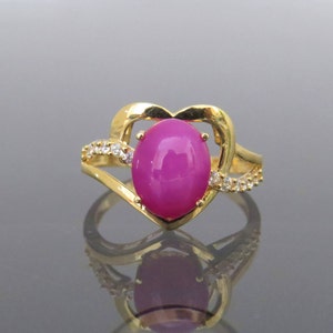 Puede incluir: Un anillo de oro con una piedra ovalada rosa engastada en un diseño en forma de corazón. El anillo está adornado con pequeñas piedras transparentes.