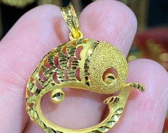May include: A gold fish pendant with a textured surface and a 24K marking. The pendant is in the shape of a fish with its mouth open and its tail curled.