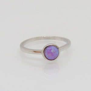 Puede incluir: Un anillo de plata con una piedra de &oacute;palo redonda de color lavanda. El &oacute;palo tiene una mezcla de tonos morados y azules. La banda del anillo es delgada y pulida, y la piedra est&aacute; engastada en un bisel simple. El anillo se muestra sobre una superficie blanca.