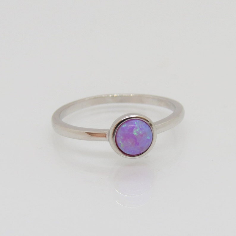 Puede incluir: Un anillo de plata con una piedra de &oacute;palo redonda de color lavanda. El &oacute;palo tiene una mezcla de tonos morados y azules. La banda del anillo es delgada y pulida, y la piedra est&aacute; engastada en un bisel simple. El anillo se muestra sobre una superficie blanca.