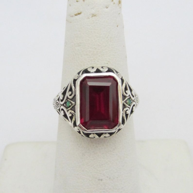 May include: A silver ring with a large, red, emerald-cut gemstone surrounded by a filigree design. Two smaller green gemstones are set on either side of the main stone.