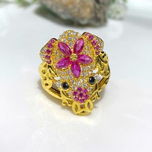Può includere: Un anello d'oro con un designo a forma di fiore rosa e pietre preziose trasparenti e rosa. L'anello ha la forma di una testa di maiale.