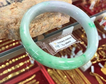 May include: A light green jade bangle with white and green swirls. The bangle is on display in a glass case with a price tag that reads "NATURAL JADITE No. 109479".