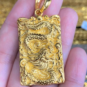 Pu&ograve; includere: Un ciondolo d'oro con un designo di drago dettagliato. Il ciondolo &egrave; rettangolare e ha un anello nella parte superiore per attaccarlo a una catena.