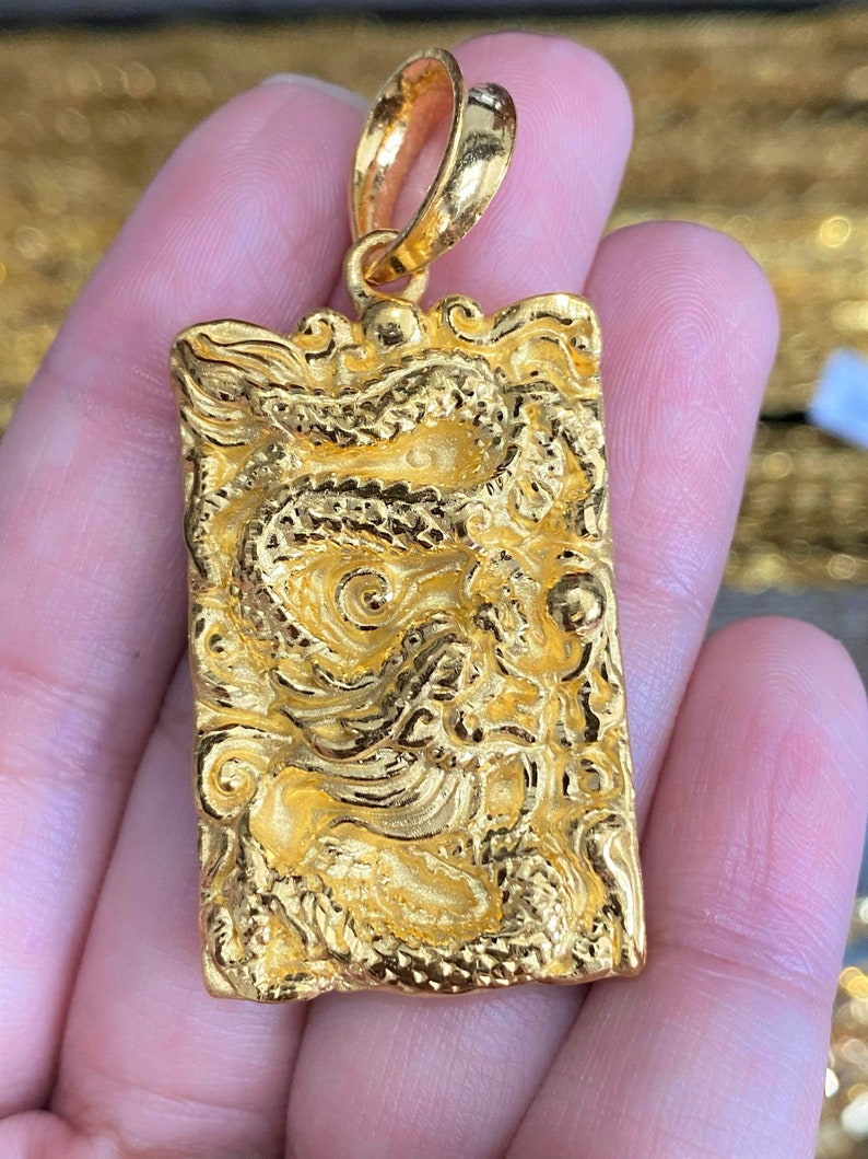 Pu&ograve; includere: Un ciondolo d'oro con un designo di drago dettagliato. Il ciondolo &egrave; rettangolare e ha un anello nella parte superiore per attaccarlo a una catena.