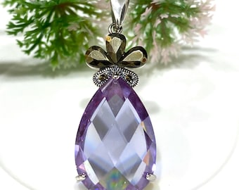 Può includere: Un ciondolo a forma di pera con una pietra preziosa viola sfaccettata e una montatura in argento. Il ciondolo è impreziosito da una corona di piccole pietre dorate.
