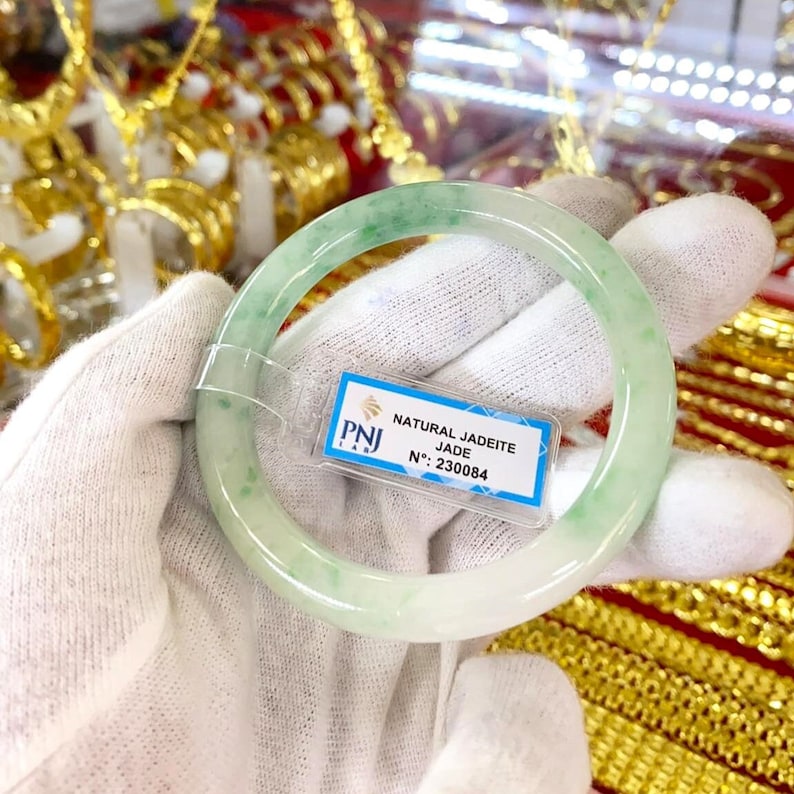 May include: A light green jadeite jade bangle bracelet with a white background. The bracelet has a label that reads "NATURAL JADEITE JADE N&deg; 230084".