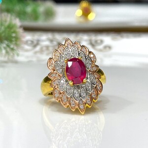 May include: A gold ring with a large oval ruby gemstone surrounded by smaller diamonds. The ring has a floral design with three layers of petals.