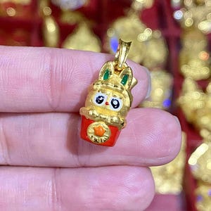 May include: A gold-colored pendant featuring a cartoon bunny in a red cup. The bunny has green ears, black eyes, and a gold crown. The cup has an orange emblem. The pendant is likely a charm or jewelry piece.