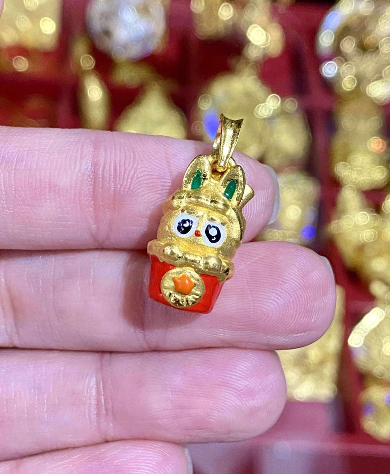 May include: A gold-colored pendant featuring a cartoon bunny in a red cup. The bunny has green ears, black eyes, and a gold crown. The cup has an orange emblem. The pendant is likely a charm or jewelry piece.