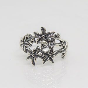 Può includere: Un anello d'argento con un designo floreale. L'anello presenta cinque fiori con cinque petali ciascuno, collegati da uno stelo delicato.