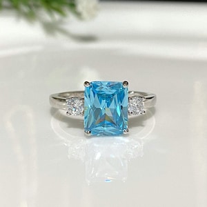 May include: A silver ring with a large, rectangular, light blue gemstone surrounded by two smaller, round, clear gemstones.