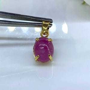 Puede incluir: Un colgante de piedra preciosa rosa con engaste de oro. El colgante tiene forma ovalada y una superficie lisa y pulida.