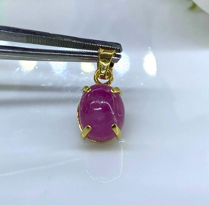 Puede incluir: Un colgante de piedra preciosa rosa con engaste de oro. El colgante tiene forma ovalada y una superficie lisa y pulida.