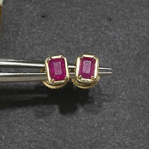 May include: A pair of gold earrings with red ruby gemstones. The earrings have a square shape and are set in a gold frame.