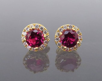 May include: A pair of gold earrings with a red gemstone surrounded by a halo of smaller white gemstones.