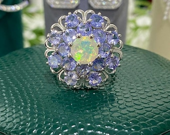 Può includere: Un anello d'argento con un'opale centrale circondata da piccole pietre rotonde viola. L'anello è posto su una superficie verde strutturata, con orecchini di diamanti sullo sfondo.