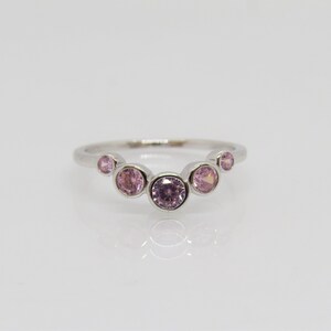 May include: A silver ring featuring a series of pink gemstones. The gemstones are arranged in a curved pattern across the top of the ring. The ring has a polished, reflective surface, and the gemstones vary slightly in size and shade of pink.