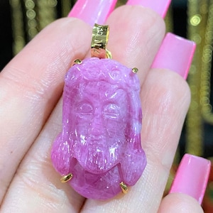 Puede incluir: Un colgante de piedra preciosa tallada de color rosa con un rostro detallado y detalles dorados. El colgante se sostiene en una mano con esmalte de uñas rosa.
