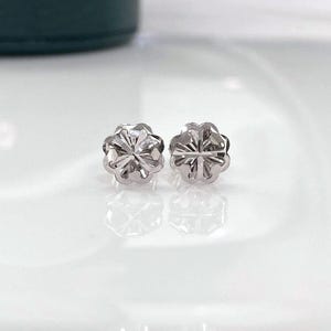 Puede incluir: Un par de peque&ntilde;os pendientes de bot&oacute;n plateados. Cada pendiente tiene un dise&ntilde;o floral con m&uacute;ltiples p&eacute;talos y una superficie texturizada. Los pendientes se muestran sobre una superficie blanca reflectante.