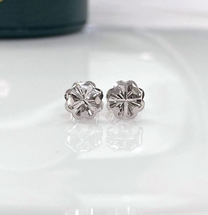 Puede incluir: Un par de peque&ntilde;os pendientes de bot&oacute;n plateados. Cada pendiente tiene un dise&ntilde;o floral con m&uacute;ltiples p&eacute;talos y una superficie texturizada. Los pendientes se muestran sobre una superficie blanca reflectante.