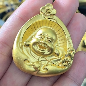 May include: A gold-colored pendant featuring a smiling Buddha figure with a flower design. The pendant is in the shape of a teardrop and has intricate details.