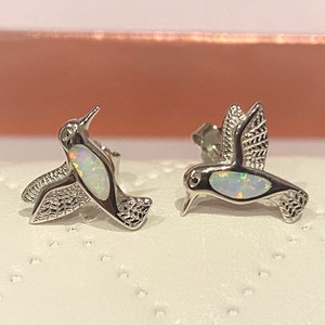 May include: A pair of silver hummingbird earrings with opal accents. The hummingbirds are facing opposite directions.
