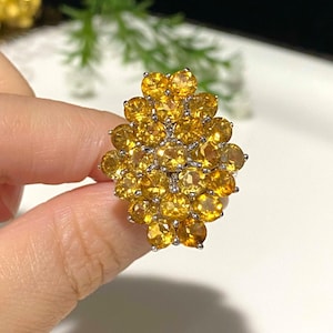 May include: A large, yellow gemstone ring with a cluster of round stones set in silver. The ring is designed in a flower shape.
