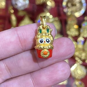 May include: A gold-colored pendant in the shape of a cartoon bunny with green accents on the ears. The bunny has large black eyes and is set on a red base with a gold star. The pendant is likely a charm or jewelry item.