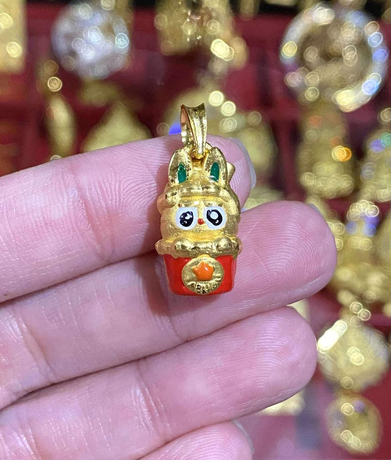 May include: A gold-colored pendant in the shape of a cartoon bunny with green accents on the ears. The bunny has large black eyes and is set on a red base with a gold star. The pendant is likely a charm or jewelry item.
