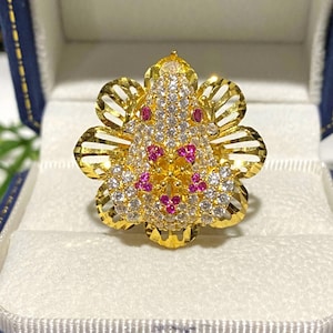 Peut inclure: Une bague en or avec un motif de fleur. Le centre de la fleur est un groupe de petites pierres blanches et roses. La bague est dans un écrin en velours bleu.