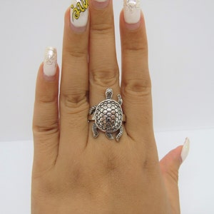 May include: A silver ring featuring a detailed turtle design. The turtle's shell is textured and the ring is worn on a hand with painted fingernails.