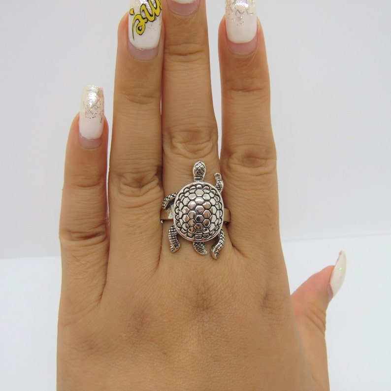 May include: A silver ring featuring a detailed turtle design. The turtle's shell is textured and the ring is worn on a hand with painted fingernails.