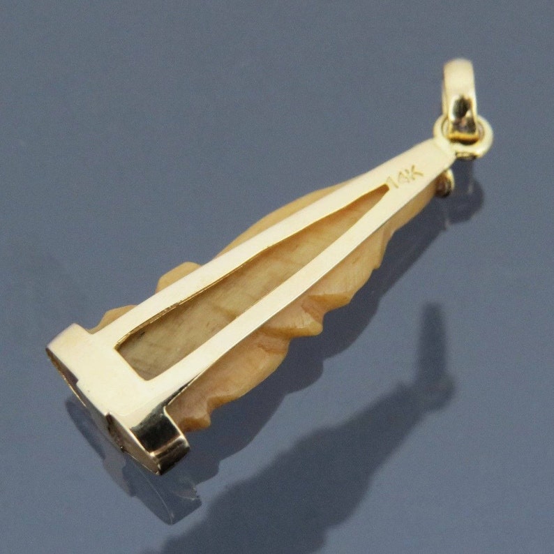 May include: A gold-colored pendant with a carved, light-tan figure. The pendant has a rectangular base and a loop at the top for a chain. The inscription "14K" is visible on the pendant. The pendant is likely made of gold.