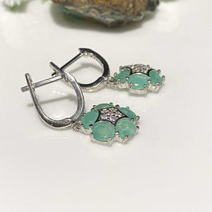 May include: A pair of silver earrings with a lever back closure. Each earring features a cluster of light green gemstones surrounded by smaller clear stones. The earrings are displayed on a white surface.