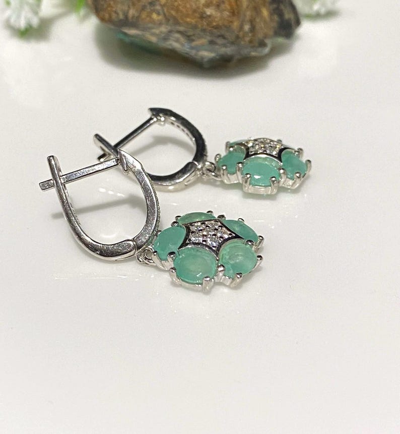 May include: A pair of silver earrings with a lever back closure. Each earring features a cluster of light green gemstones surrounded by smaller clear stones. The earrings are displayed on a white surface.