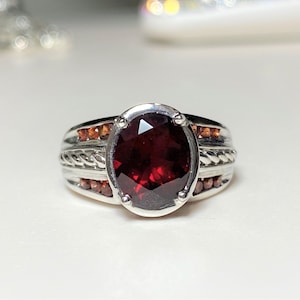 May include: A silver ring with a large, oval-shaped, red gemstone surrounded by smaller, orange gemstones.