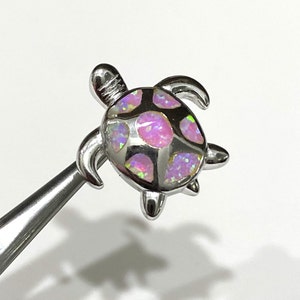 May include: A silver turtle pendant with a pink and white opal inlay. The turtle is facing right and has a detailed shell.