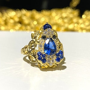 May include: A gold-toned ring featuring a large, pear-shaped blue gemstone at its center, surrounded by smaller blue and clear stones. The ring has an ornate, detailed design with a floral or leaf-like pattern.