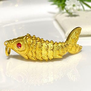 Puede incluir: Un amuleto de pez de color dorado con un ojo de piedra preciosa roja. El pez est&aacute; detallado con escamas y aletas.