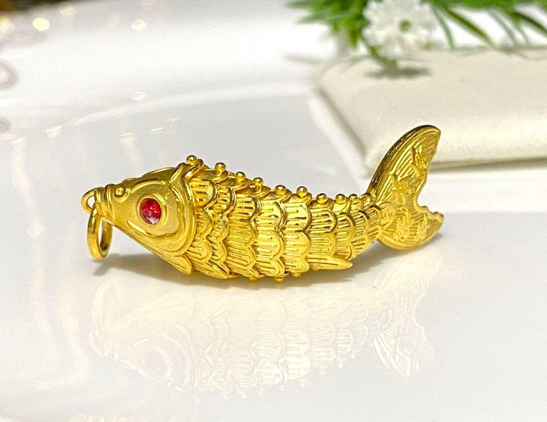 Puede incluir: Un amuleto de pez de color dorado con un ojo de piedra preciosa roja. El pez est&aacute; detallado con escamas y aletas.