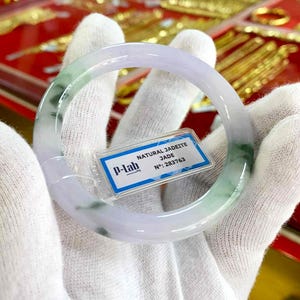 May include: A jadeite jade bangle bracelet with a translucent appearance, displaying green and lavender tones. The bracelet is circular and polished, with a tag stating "NATURAL JADEITE JADE" and a serial number. Gold jewellery is visible in the blurred background.