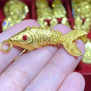 May include: A gold-colored fish charm with a red gemstone eye. The charm has a loop for attaching to a necklace or bracelet.