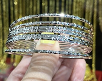 May include: A set of six thin, silver-colored metal bangles featuring a repeating geometric diamond-cut pattern along the exterior surface. Each bangle has a polished, reflective finish that catches the light, creating a bright, metallic appearance.