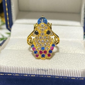 Peut inclure: Une bague en or en forme de grenouille avec des pierres précieuses bleues, roses et blanches. La grenouille porte une couronne avec une pierre précieuse bleue sur le dessus.