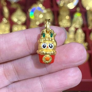 May include: A gold-colored pendant featuring a cartoon rabbit design with green accents on the ears. The rabbit is atop a red cup with an orange emblem. The pendant is held in a hand, with other gold items in the background.
