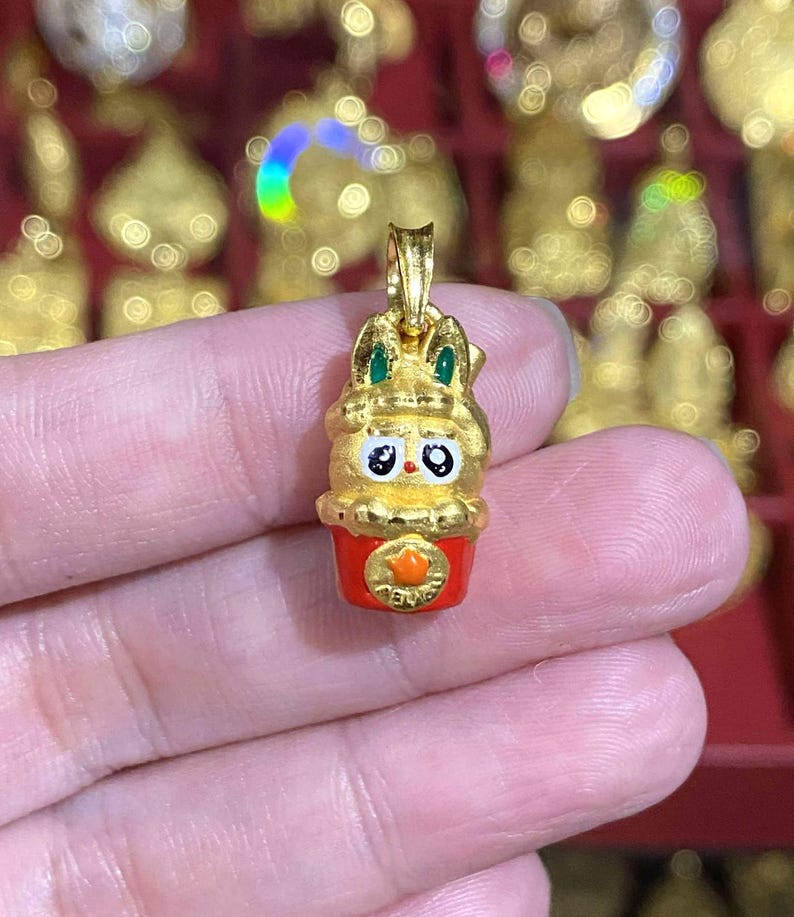 May include: A gold-colored pendant featuring a cartoon rabbit design with green accents on the ears. The rabbit is atop a red cup with an orange emblem. The pendant is held in a hand, with other gold items in the background.