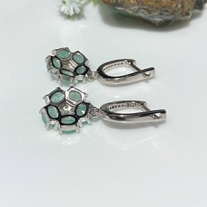May include: A pair of silver-toned drop earrings. Each earring features a cluster of oval-shaped, light green gemstones set in a floral design. The earrings have a lever-back closure and are displayed on a white surface.